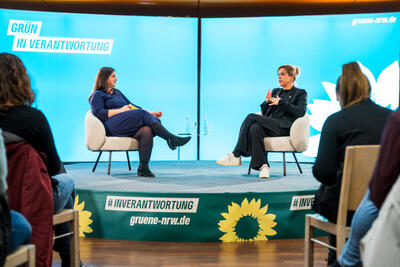 Aus der Sicht des Publikums bzw. zwischen einigen Personen hindurch sieht man zwei Frauen, die auf einer halbrunden Bühne auf Cocktail-Sesseln sitzen und miteinander im Gespräch sind. Die Bühne ist in den Farben der Grünen gestaltet und im Hintergrund mit 